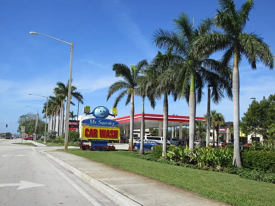 Fachada de comércio com letreiro e palmeiras na Atlantic Boulevard, em Pompano Beach, com posto de gasolina ao fundo